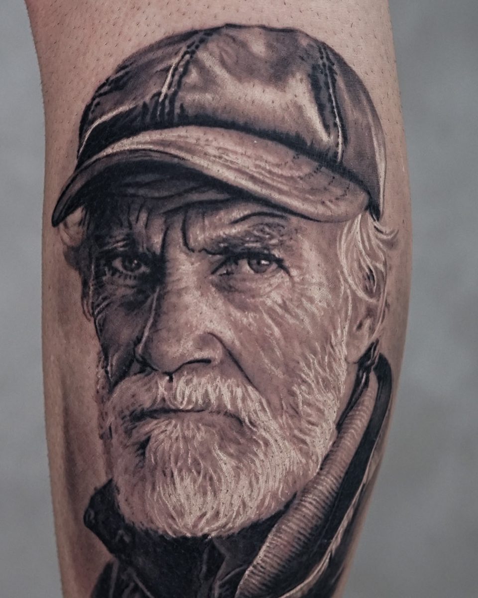 A tatuagem do curso cicatrizada, em novembro de 2024: retrato de um homem idoso de boné, em realismo preto e cinza, na panturrilha